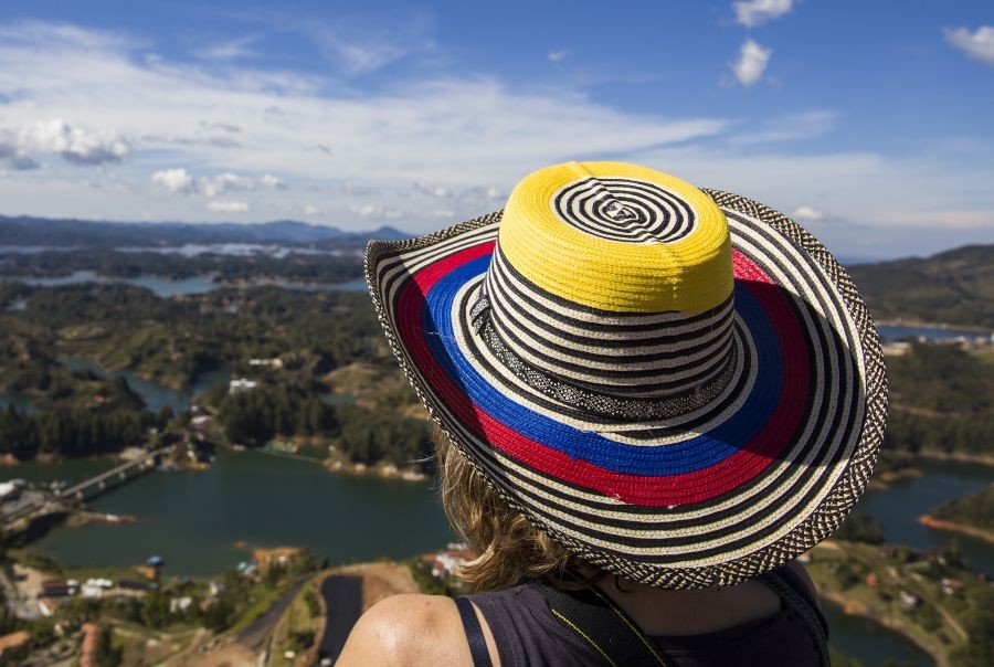 Persona de espaldas con sombrero decorado con los colores de la bandera de Colombia, contemplando un paisaje montañoso con lagos y cielo despejado